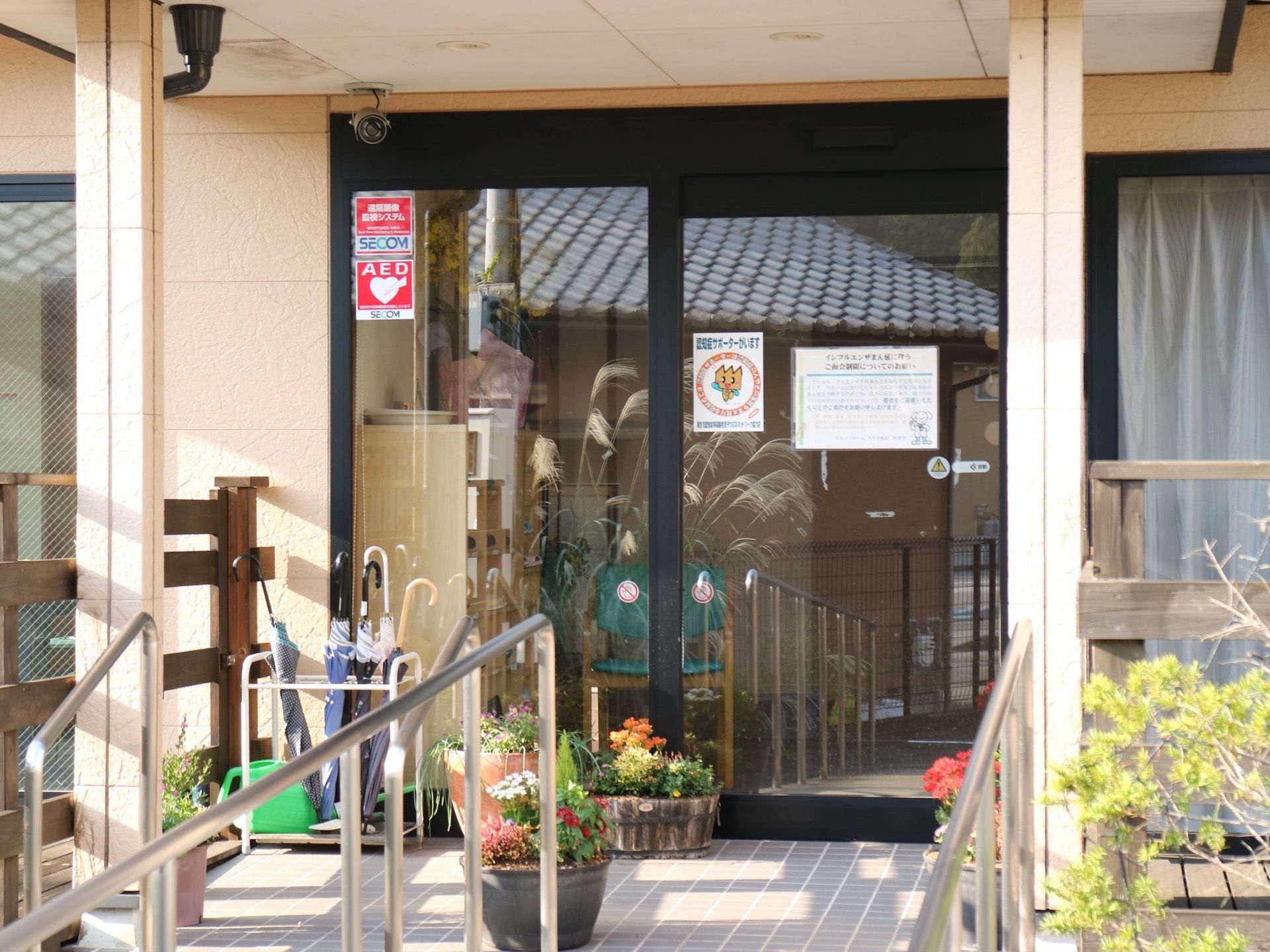 玄関の入口は自動ドアになっており、上には防犯カメラが設置されている。ドアの前には傘立てや、鉢植えの花が置かれている。