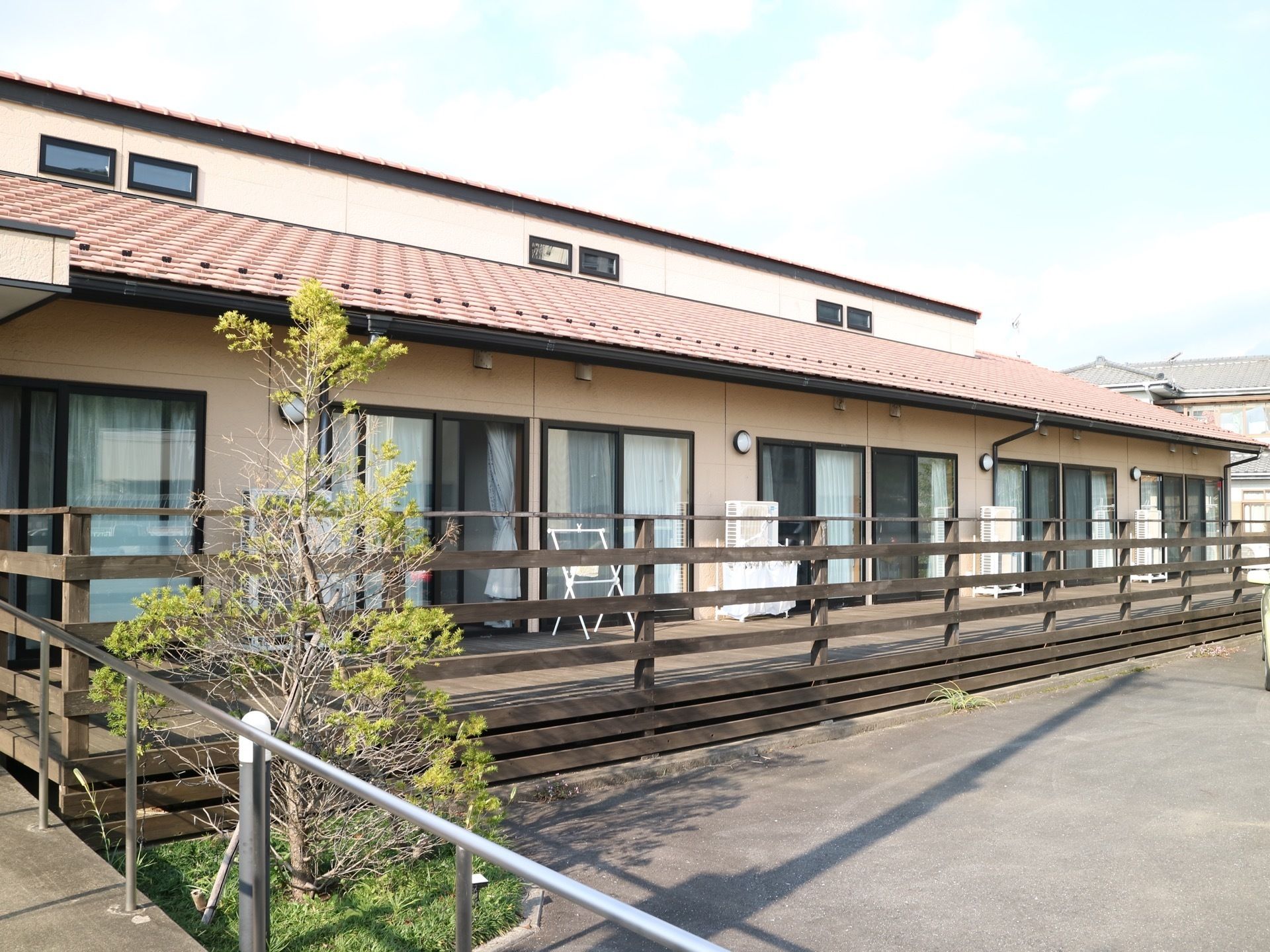 建物の横にはウッドデッキが設けられている。外側にある小さな花壇には木が植えられていて、横には手すりの付いたスロープが設置されている。