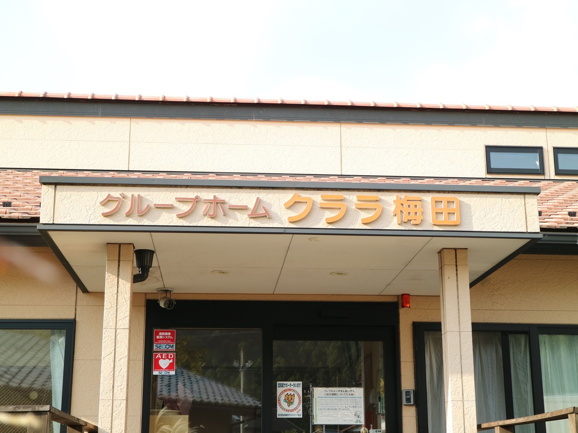 玄関の上にはひさしがあり、施設名が書かれている。自動ドアの横にはインターホンが設置されていて、ドアには張り紙やステッカーが貼られている。
