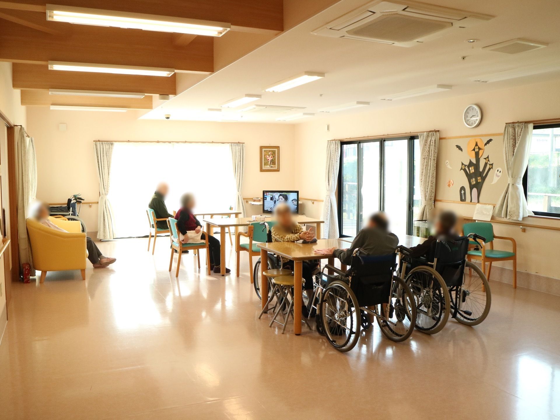 窓が多く広々とした食堂である。中央にはダイニングテーブルが２ヵ所に設置されており、左の壁際には黄色いソファが置かれている。
