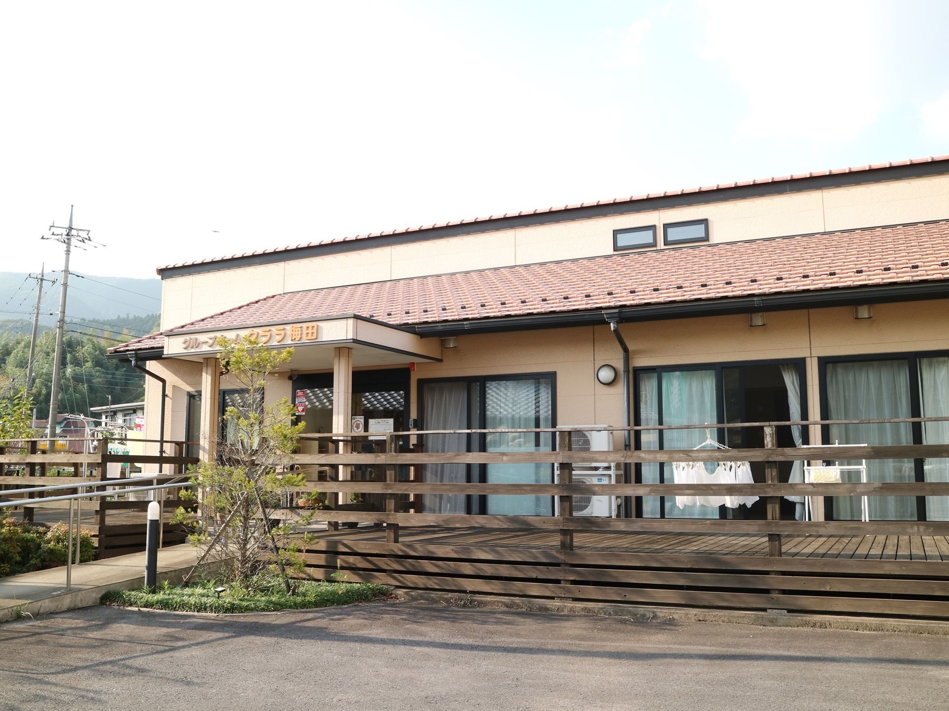 建物はベージュ色の外壁で、玄関の横には広いウッドデッキが設置されている。ウッドデッキの前には小さな花壇があり木が植えられている。