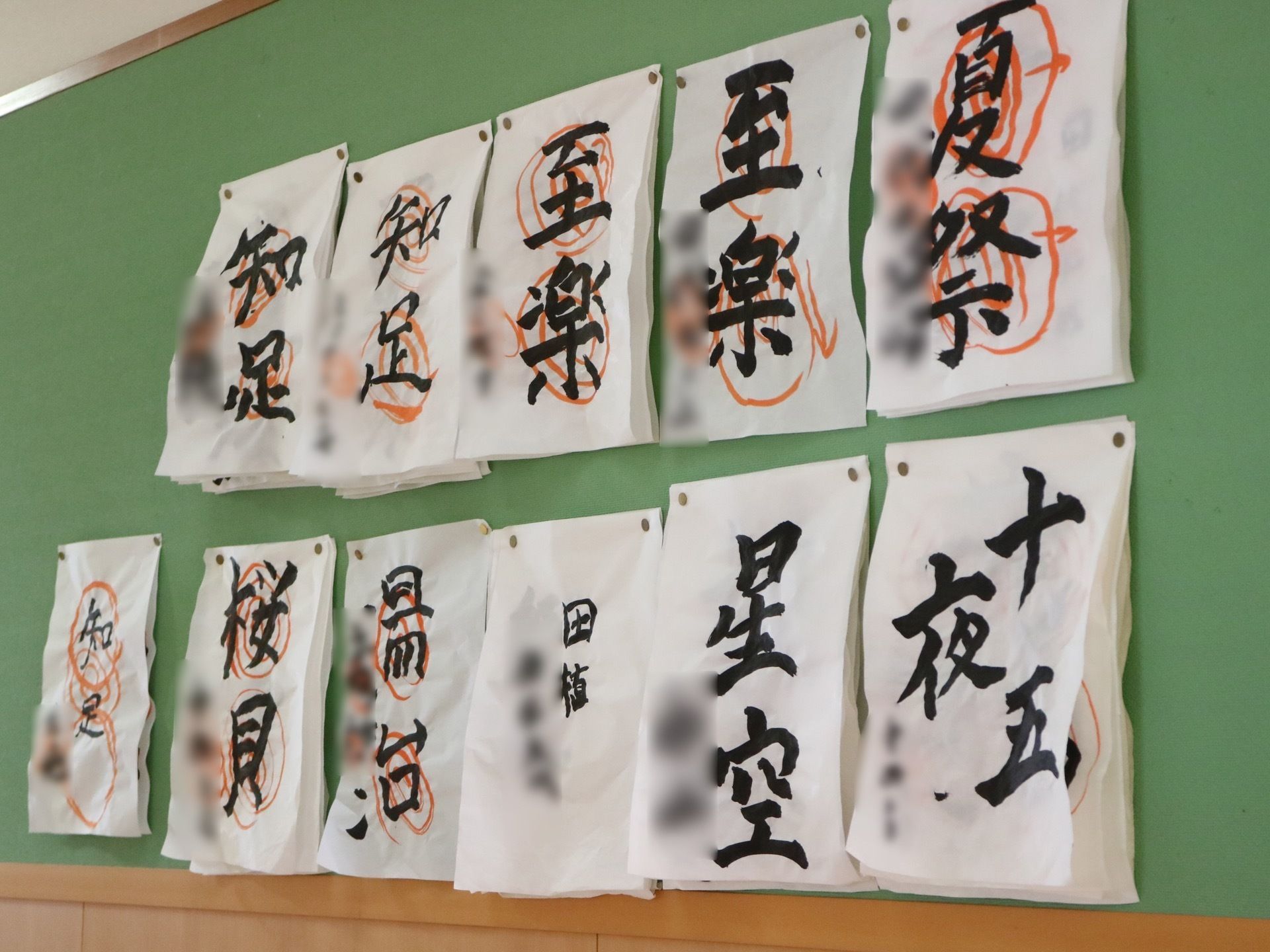 緑の掲示板に習字が貼られている。習字には赤い墨で丸が付けられており、半紙は数枚重ねて画びょうでとめられている。