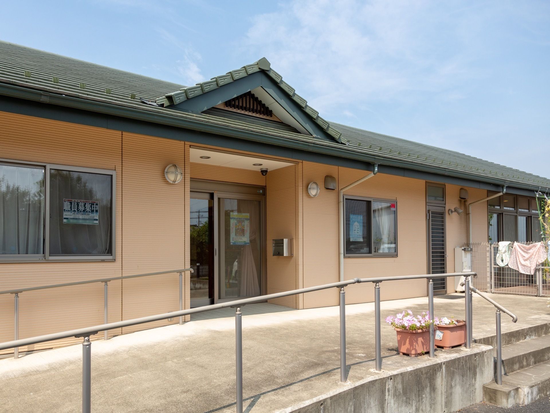 施設入口のある建物はベージュの壁面の平屋建てで、短い階段と緩やかなスロープの2通りで入口へと移動できる。スロープには手すりがついている。