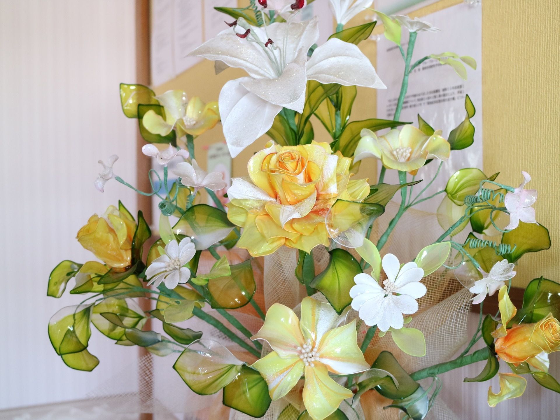 バラやゆりなど、さまざまな種類の造花が束ねられている。後ろにはベージュの掲示板があり、張り紙が貼られている。