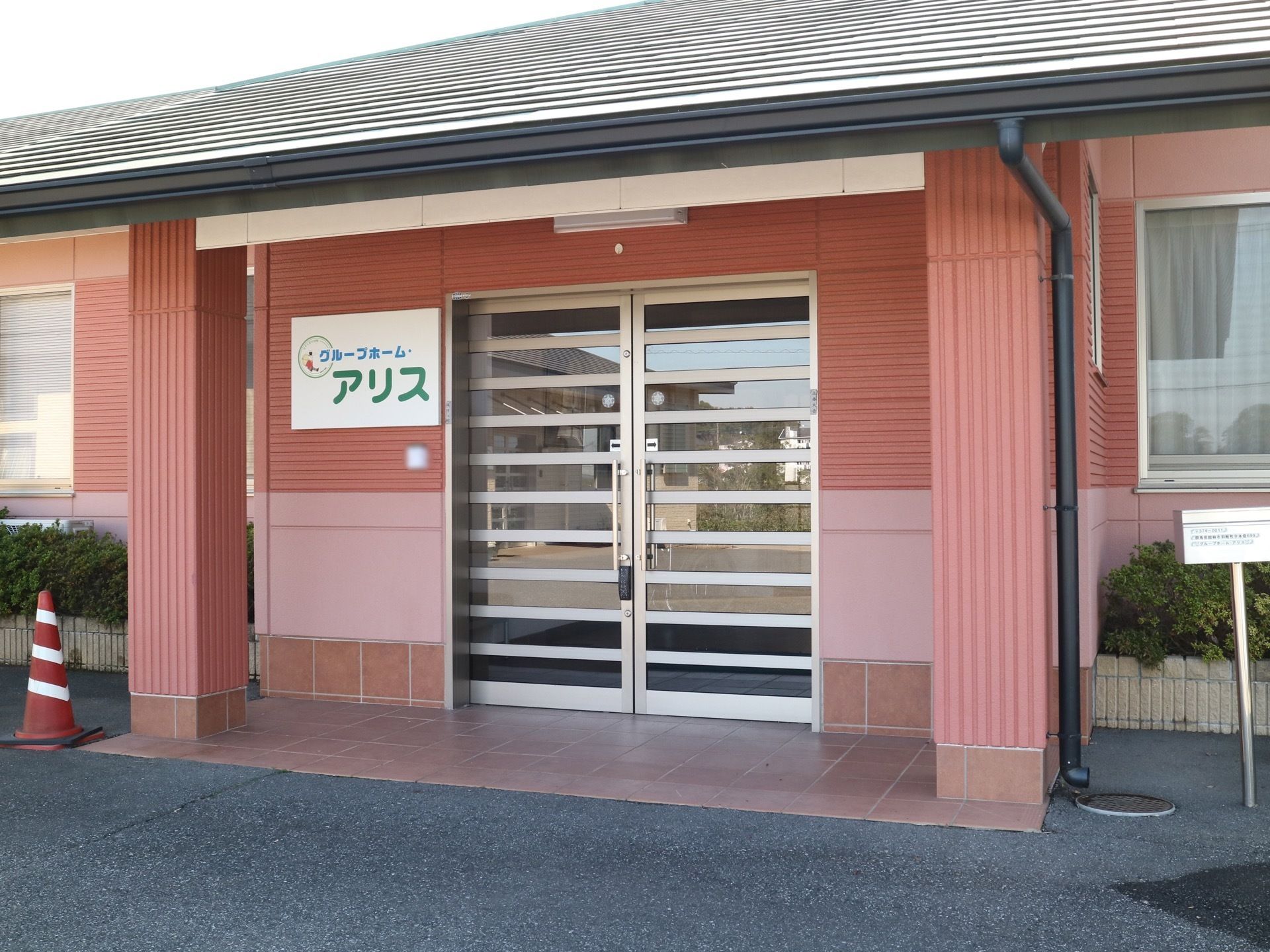 玄関はピンク色の外壁になっていて、上には屋根が付いている。入口は両開きのガラスのドアで、横の壁には看板が取り付けられている。
