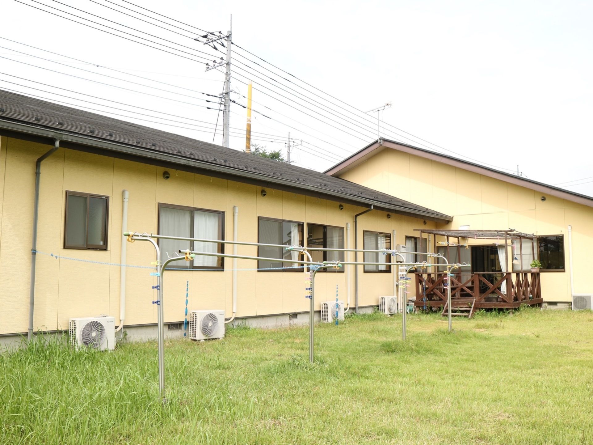 クリーム色の壁が続く長屋風の建物がある。各部屋の室外機が並んでいる。施設前は草が生えた空き地になっている。