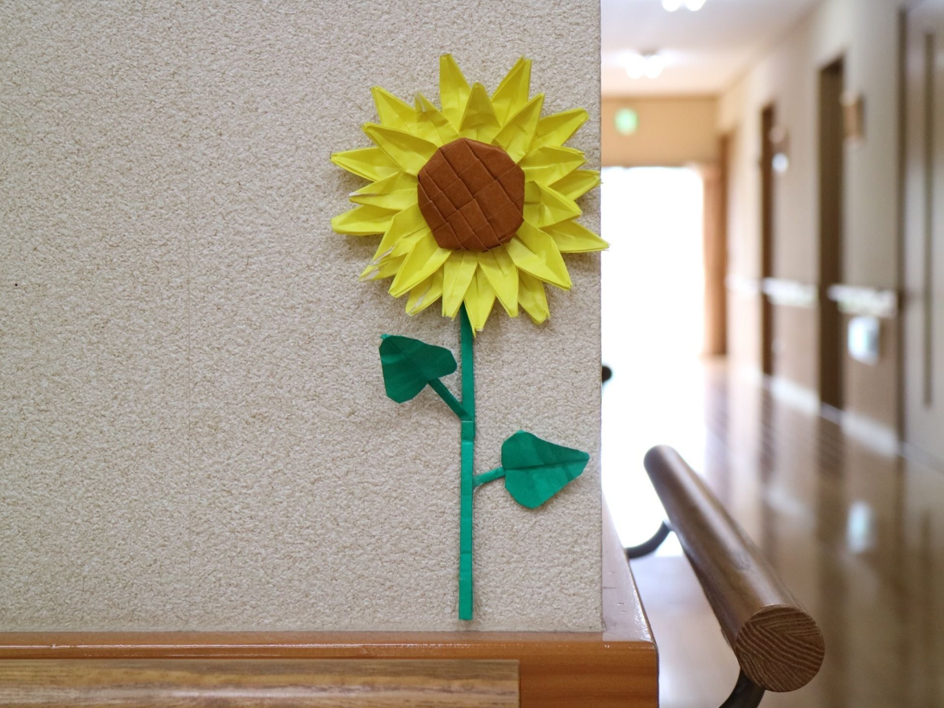 L字に曲がった廊下があり、壁に折り紙の飾り付けがされている。ひまわりの花をかたどった折り紙の作品が貼られている。
