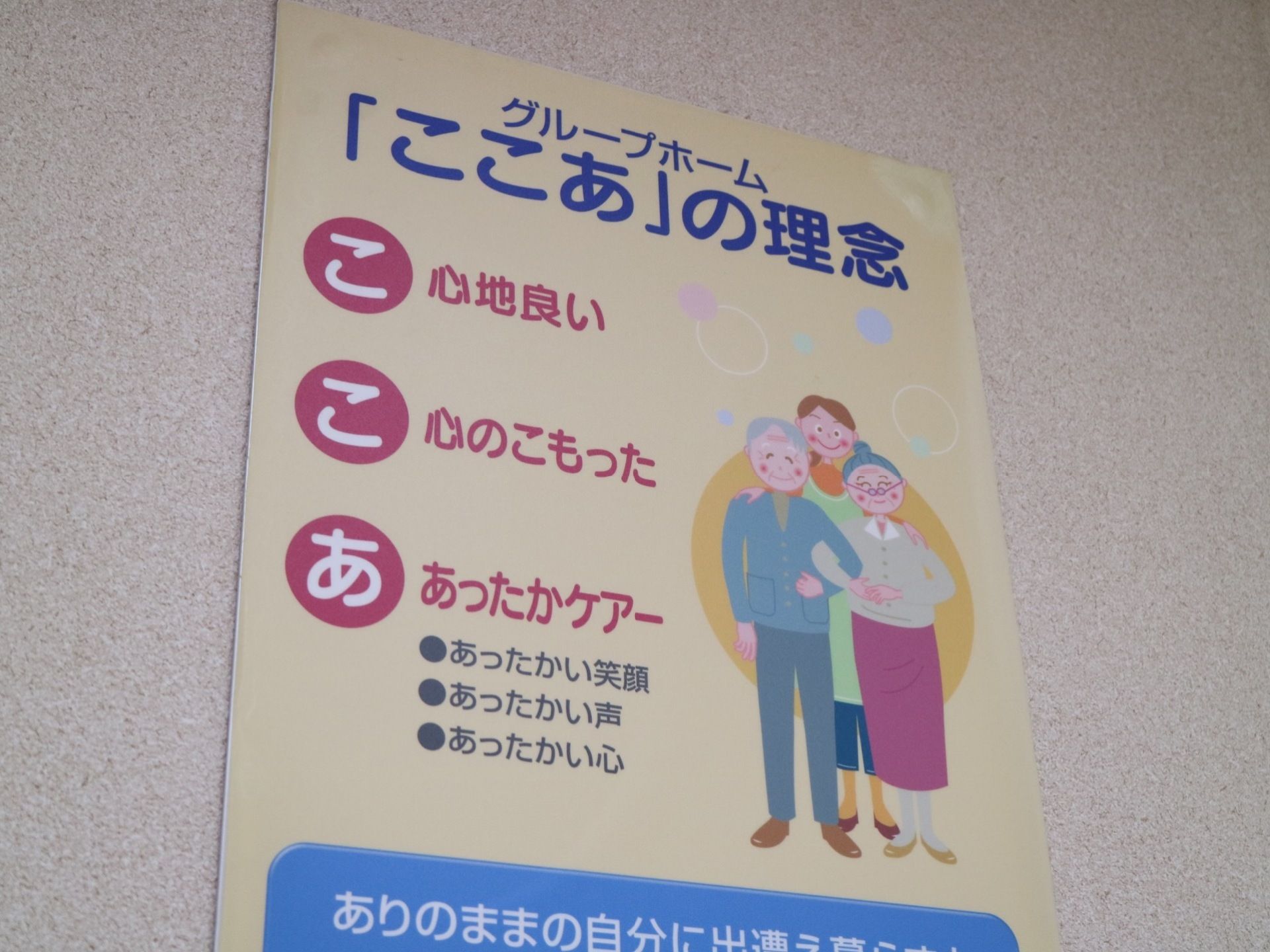 白い壁に薄い黄色の紙が貼られている。施設名をあいうえお作文にした理念が書かれている。お年寄りのイラストが描かれている。