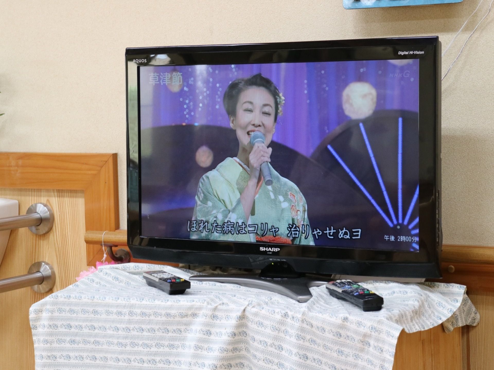 壁沿いに置かれた棚に白い布がかけられ、その上に大型液晶テレビが置かれている。テレビの前にリモコンがある。