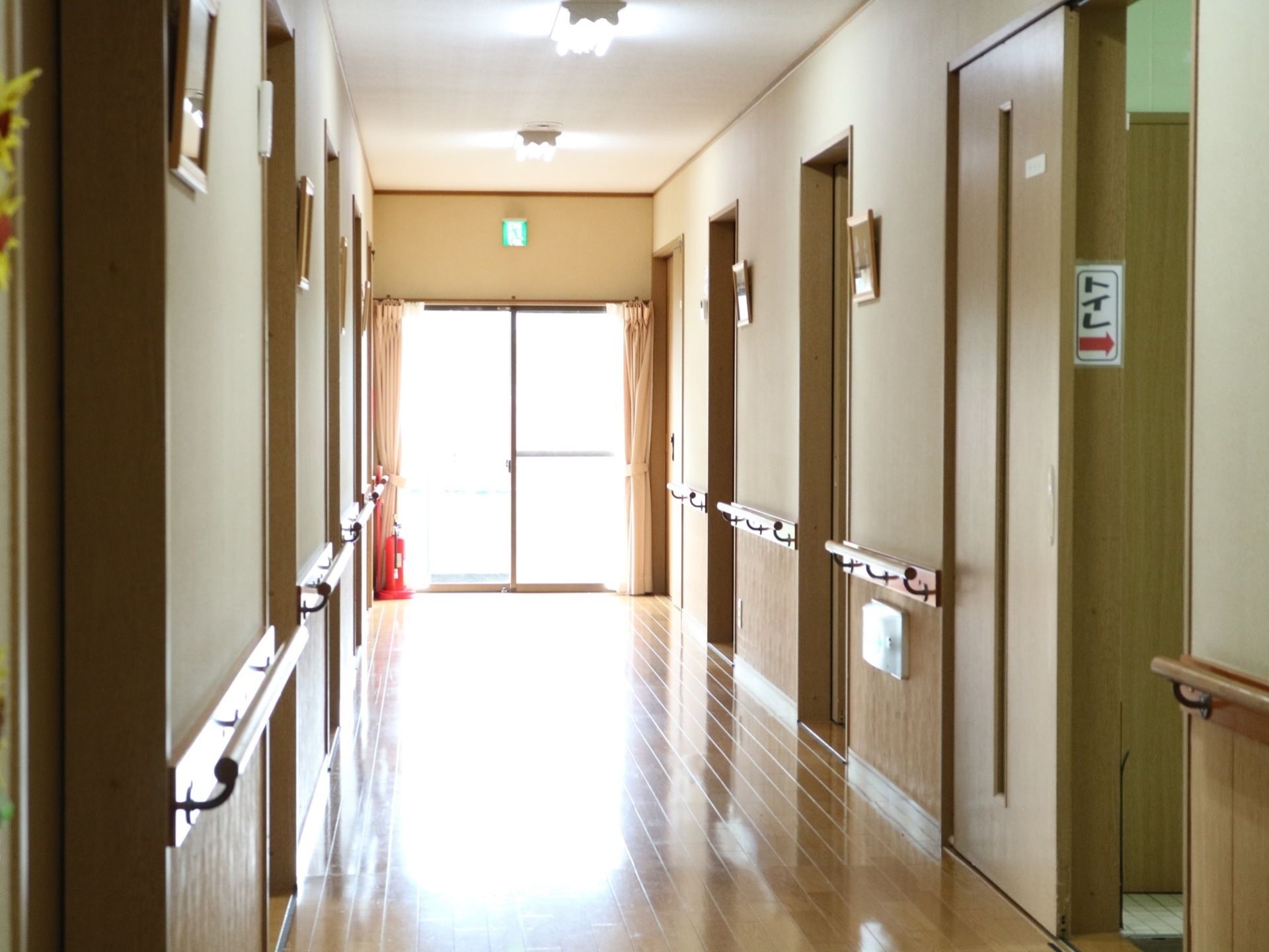 板張りの廊下の突き当たりに大きな窓がある。非常口の表示がされ、たくさんの外光が差し込んでいる。手前にトイレの入口がある。