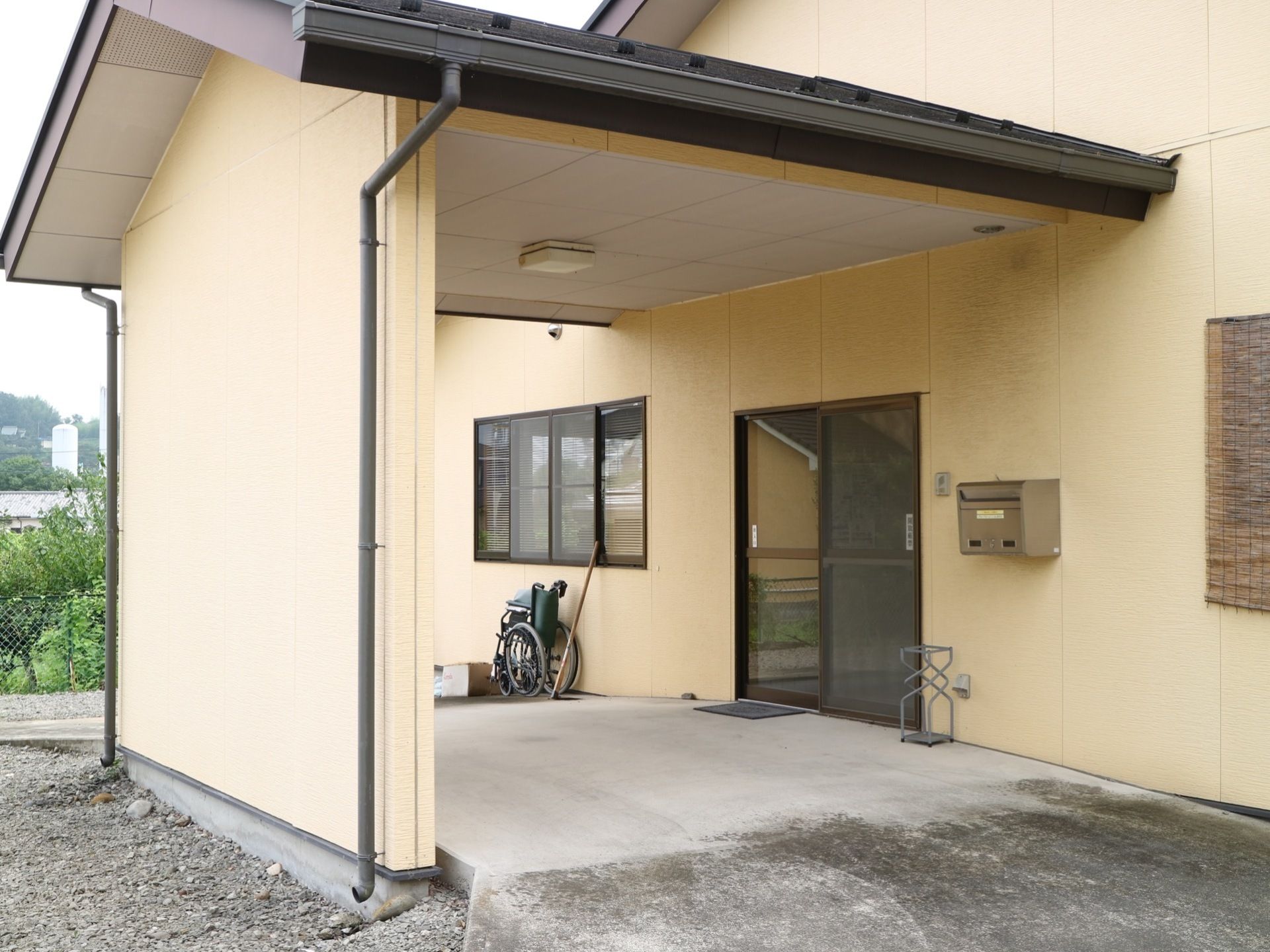 ベージュの外壁の建物で、玄関前は三角の屋根が乗っている。入口扉の横にポストがあり、折り畳みの車椅子がある。