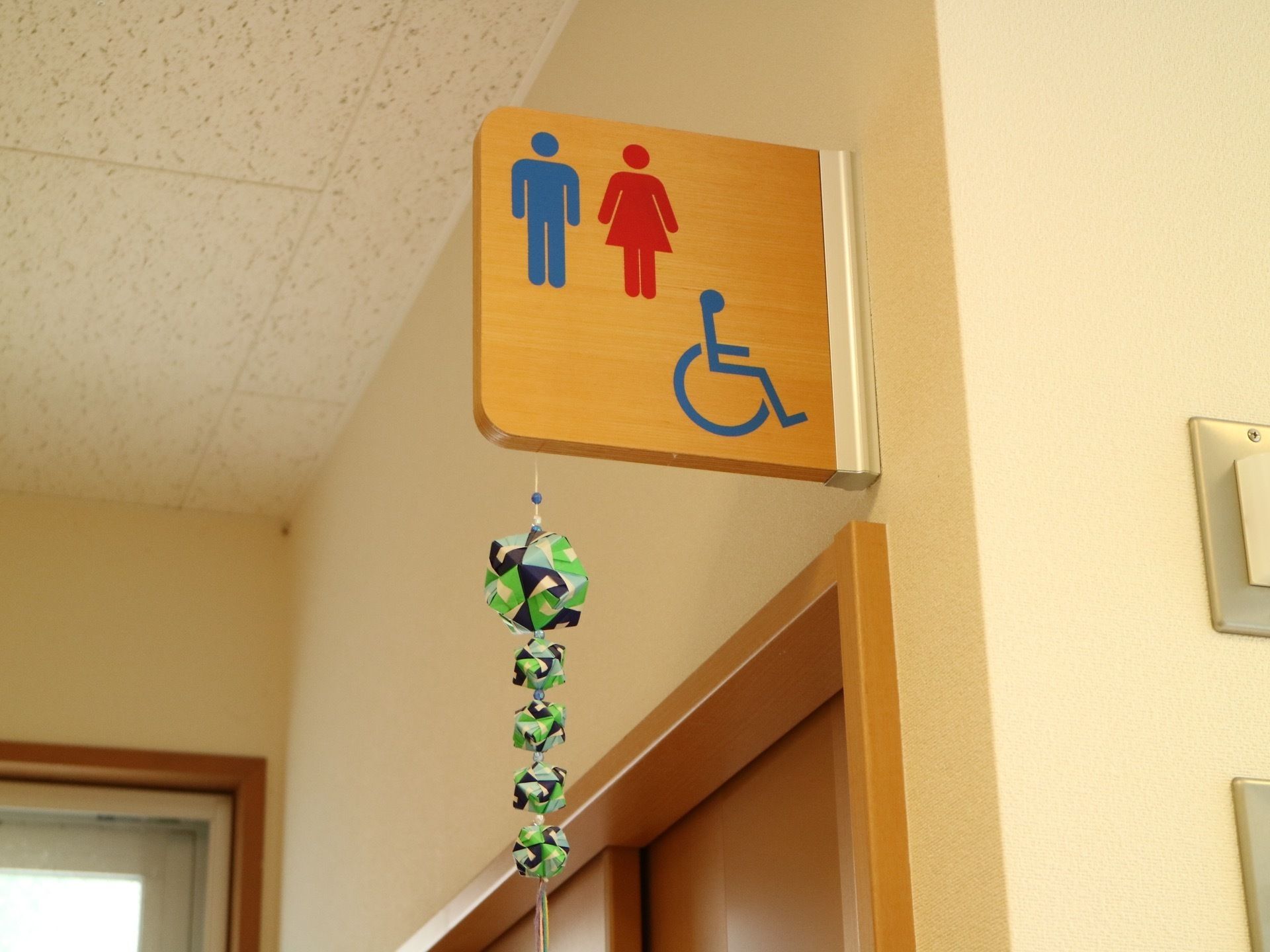 ベージュ色の壁の廊下である。天井近くにトイレの標識がかかっている。男女と車椅子のマークが描かれている。