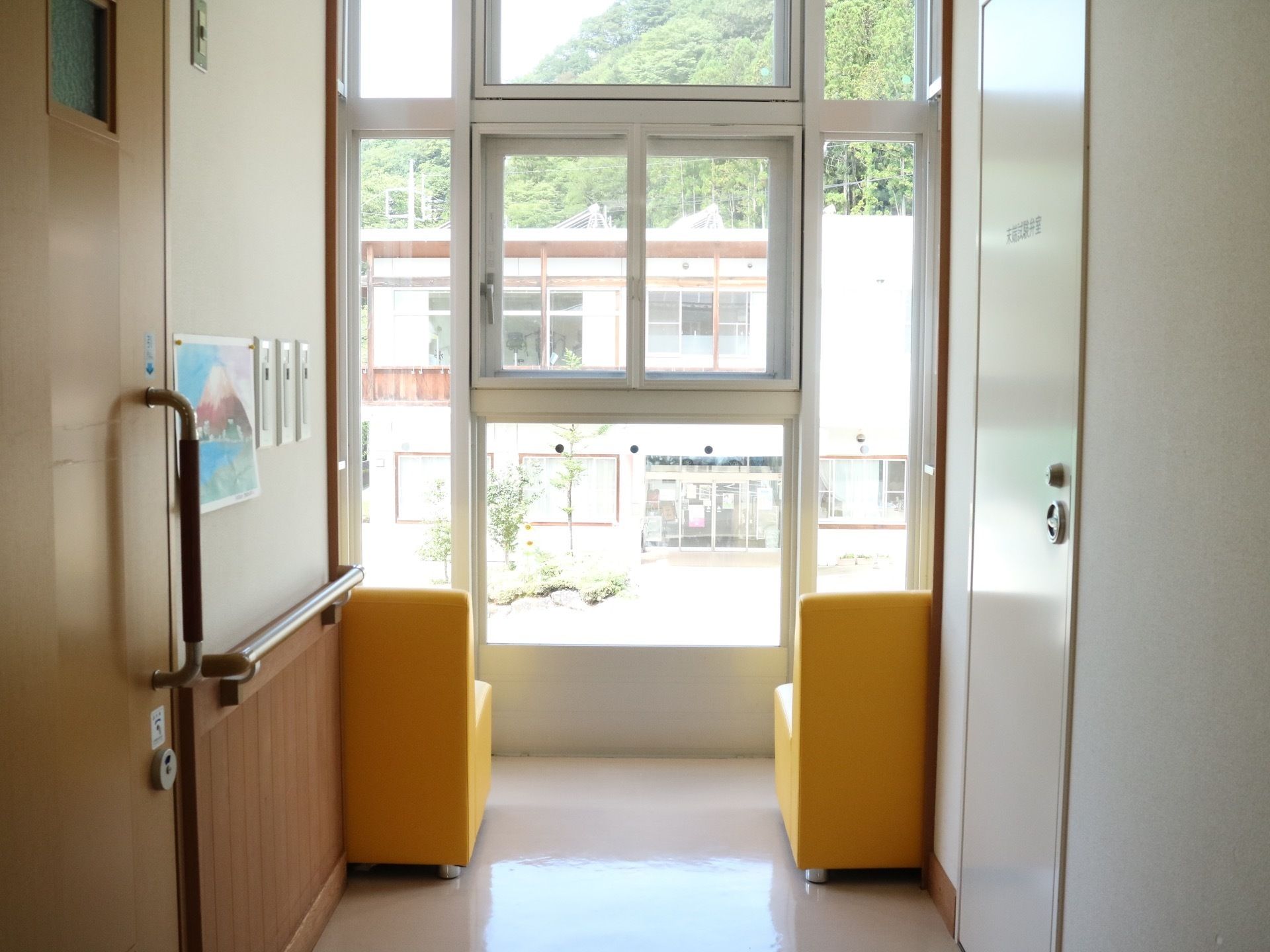 廊下の突き当りは窓があり、外の景色が見えている。壁に沿って黄色い１人掛けの椅子が２脚並べられている。