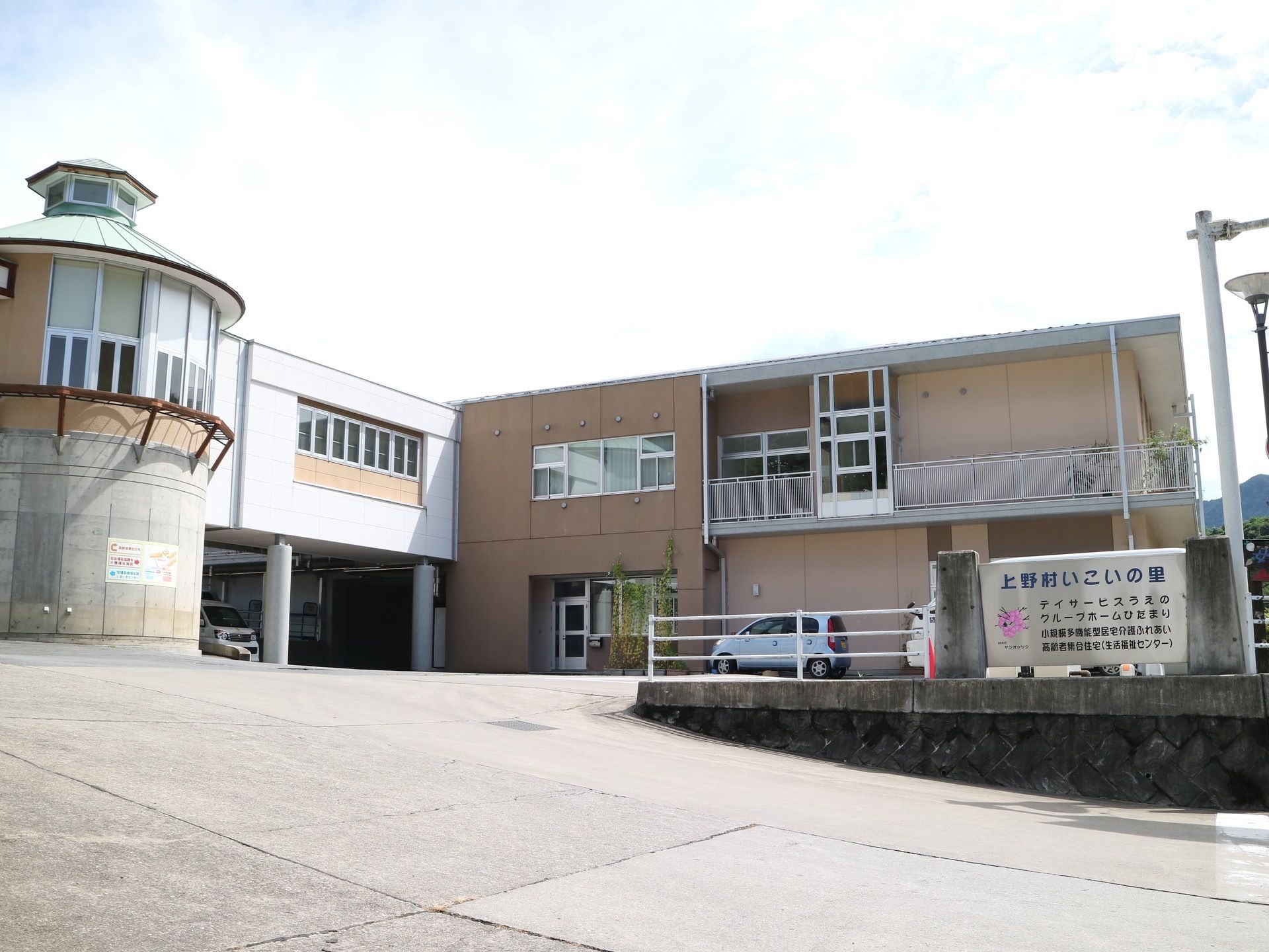 建物は２階建てで、一部が円柱型の建物になっている。施設前までは緩やかな上り坂になっている。入り口に看板が立っている。
