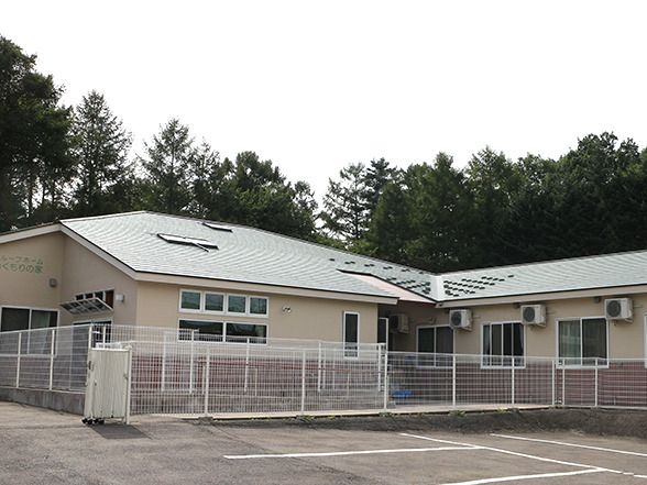 緑に囲まれた環境の中にあるライトグレー系の平屋建ての建物。敷地内には駐車場もある。