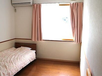 レースカーテンとカーテンが付いた大きな窓が設置された個室の部屋。寝具付きのベッドやエアコンがある。