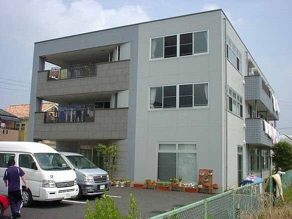 グレー系の外観の3階建ての建物の周りには花や草木が植えられ、各フロアにベランダがあり、洗濯物が干されている。
