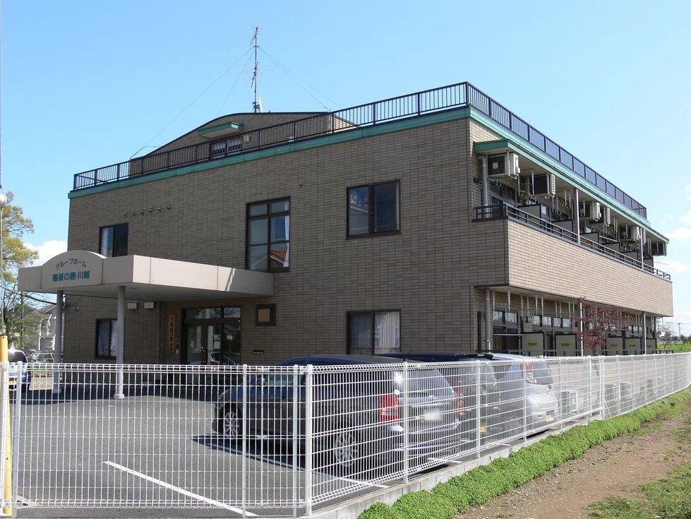 ブラウン系の２階建ての建物は日当たりのいい場所に建っていて、広い駐車場が付いている。駐車場には車が止められている。