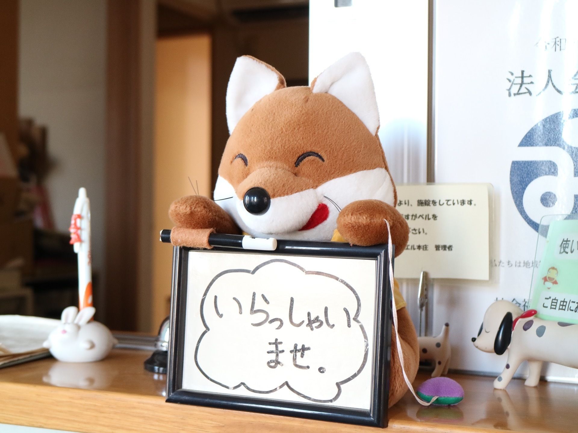 きつねのぬいぐるみが棚の上に置かれている。小さな黒い額縁を持っており、手書きでいらっしゃいませと文字が書かれている。