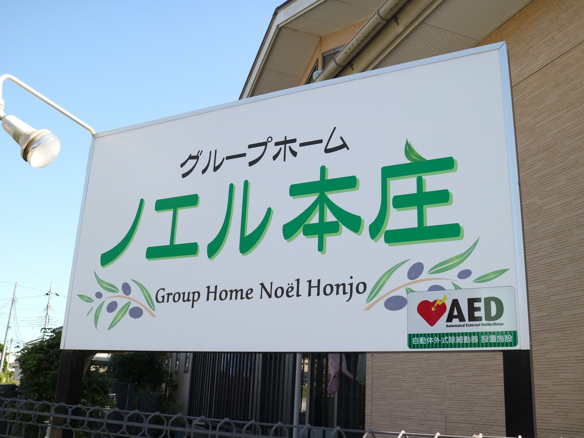 施設の入り口にある大きな看板の図である。白い板に緑系の文字で施設名が書かれ、照明が１つ取り付けられている。