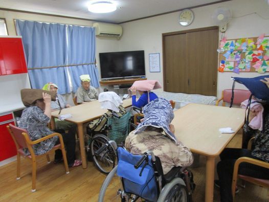 テレビがある食堂に集まって入居者たちが座布団のようなものを頭にのせて椅子に座っている様子。