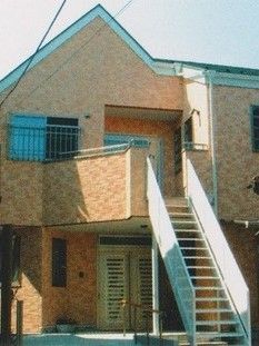 明るい日差しの下、レンガ調の２階建ての建物がある。２階部分には手すり付きの階段が付けられ、直接外に出ることができる。
