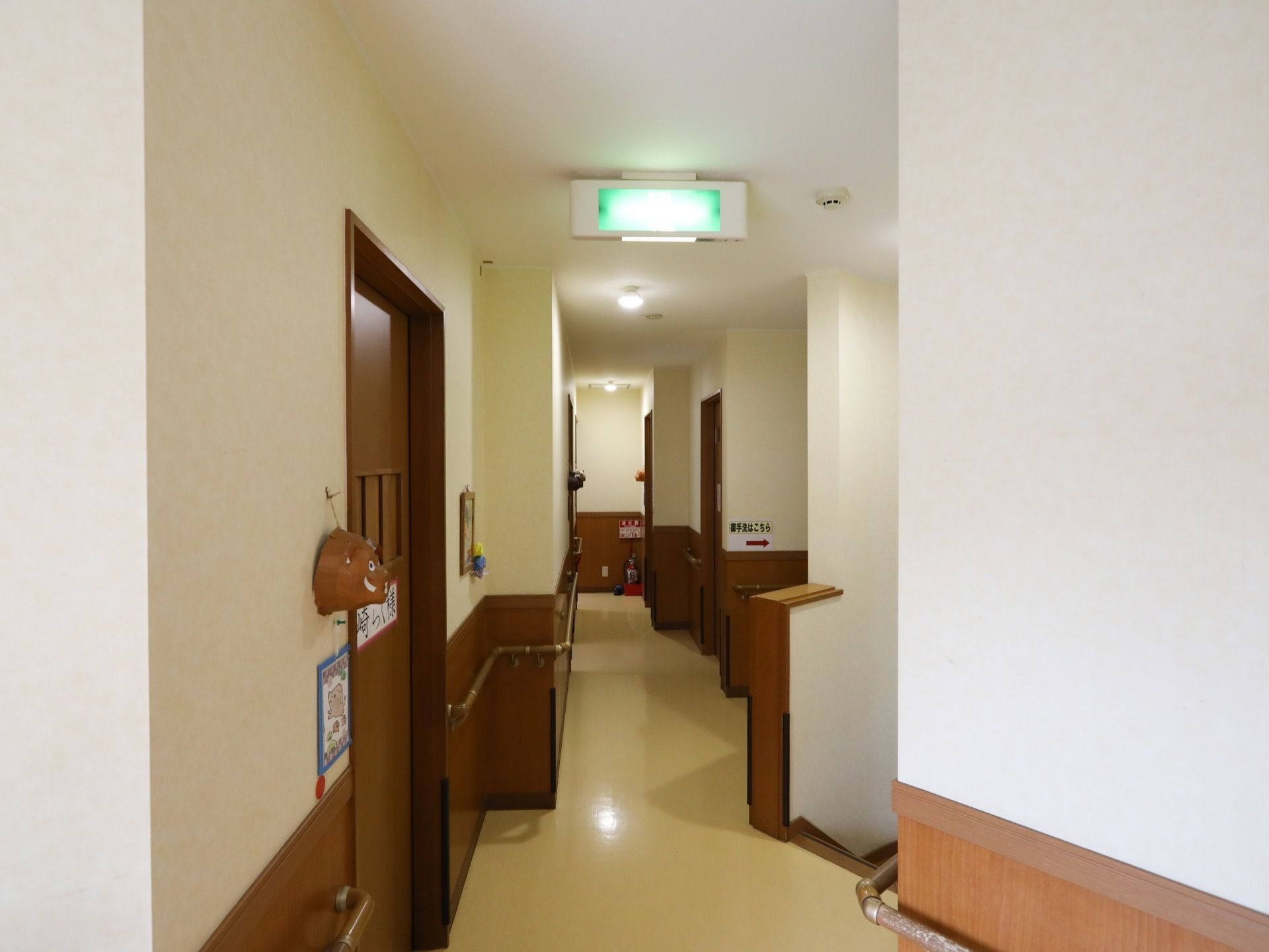 廊下の天井には非常用出口のマークが書かれている。壁には茶色い手すりが設置されている。突き当たりには消火器が置いてある。