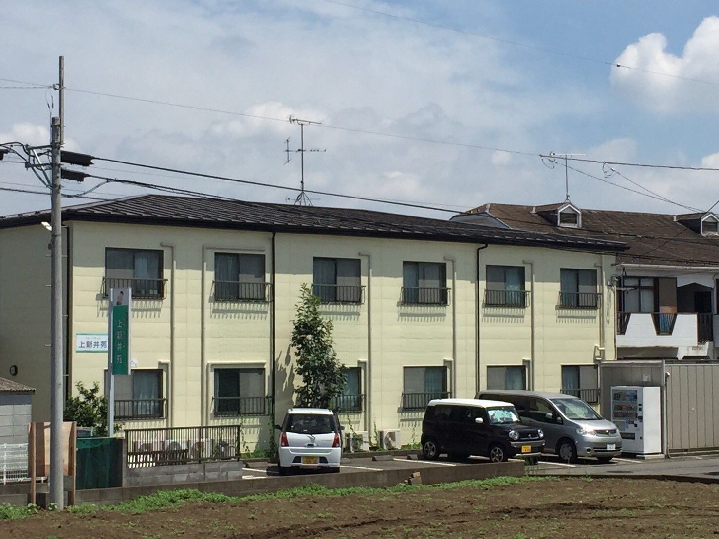２階建ての建物は日当たり良好でベージュ系の外壁。建物の前には駐車場があり、複数台の車が止まっている。