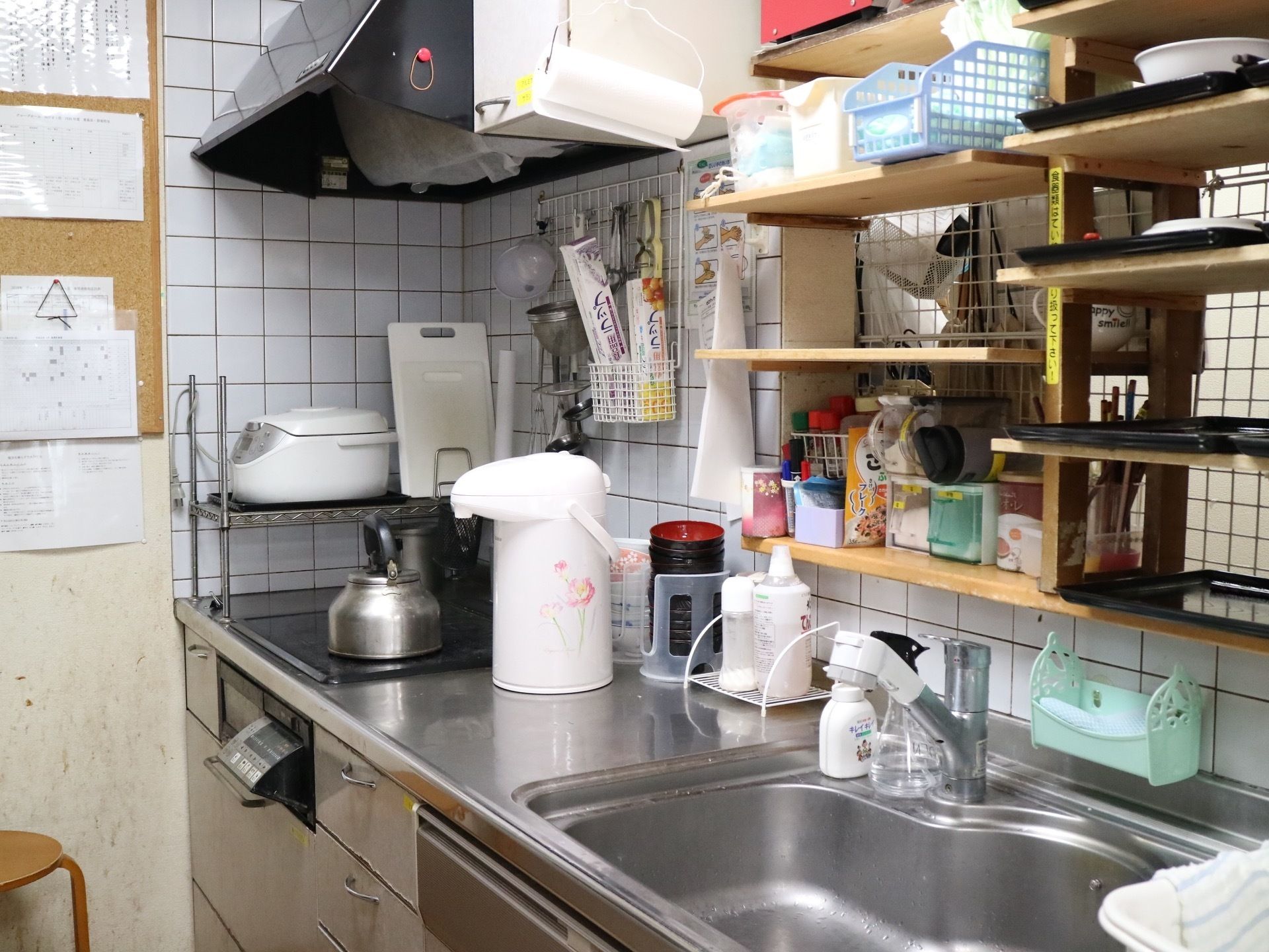 スタッフが使用するキッチンで、流し台の上に棚が作りつけられている。奥には炊飯器やポットが置いてある。