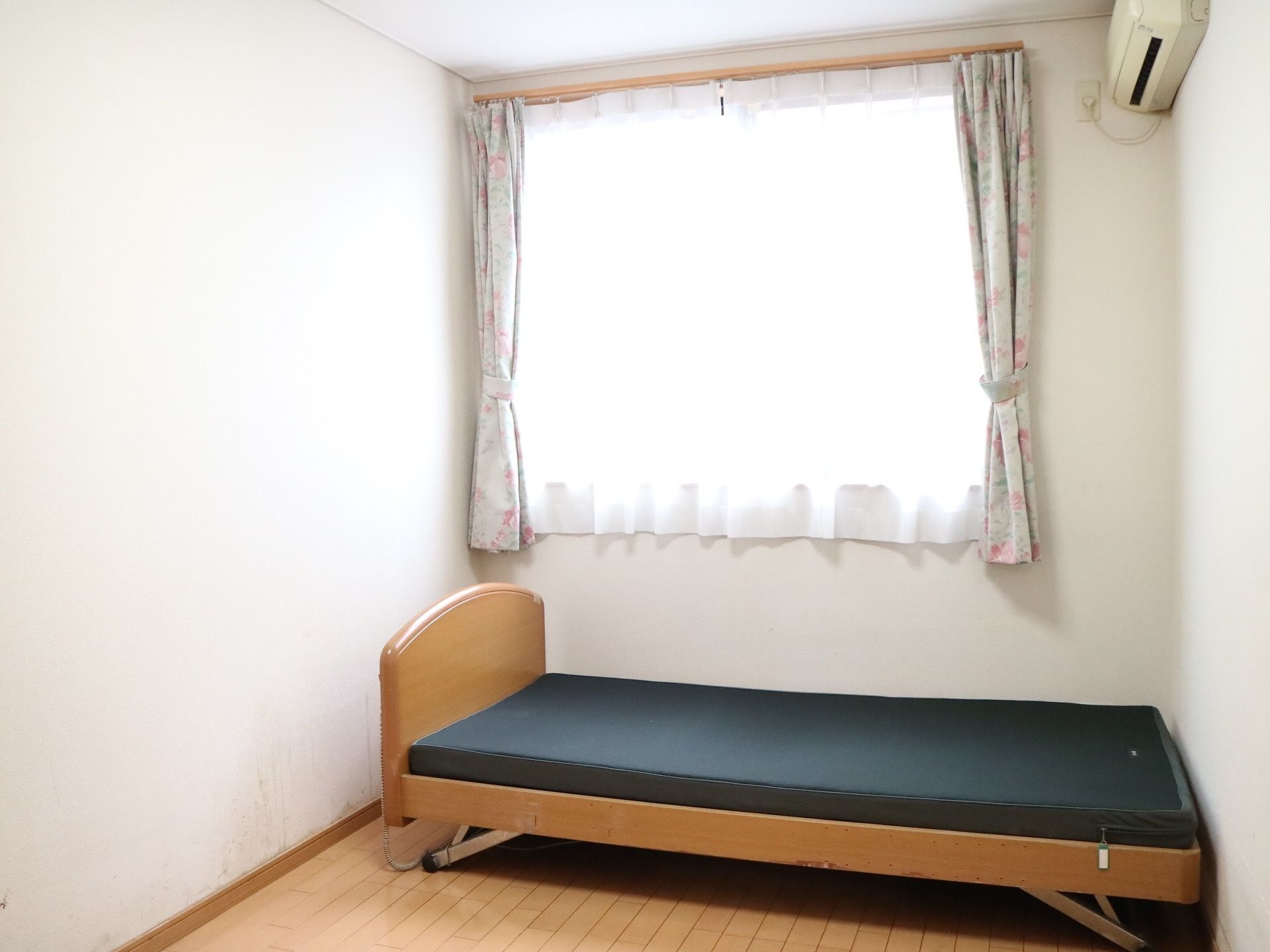 床がフローリング仕様で、白い壁に囲まれた居室である。奥に大きな窓がある。薄手のレースカーテンがかかっている。