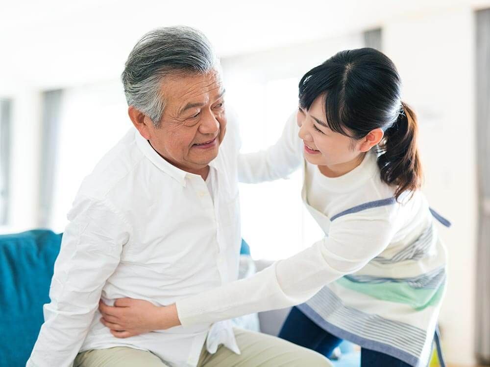 ボーダー柄のエプロンを付けた女性スタッフが白いシャツを着た高齢男性のサポートに笑顔で当たっている。