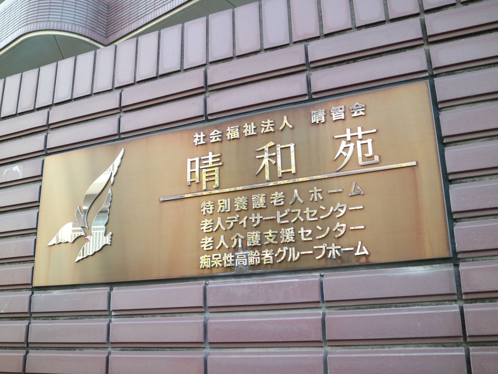 薄いピンク色の外壁の中央に大きな看板が埋め込まれている。ゴールドの板に、シルバーの素材で施設名が書かれている。
