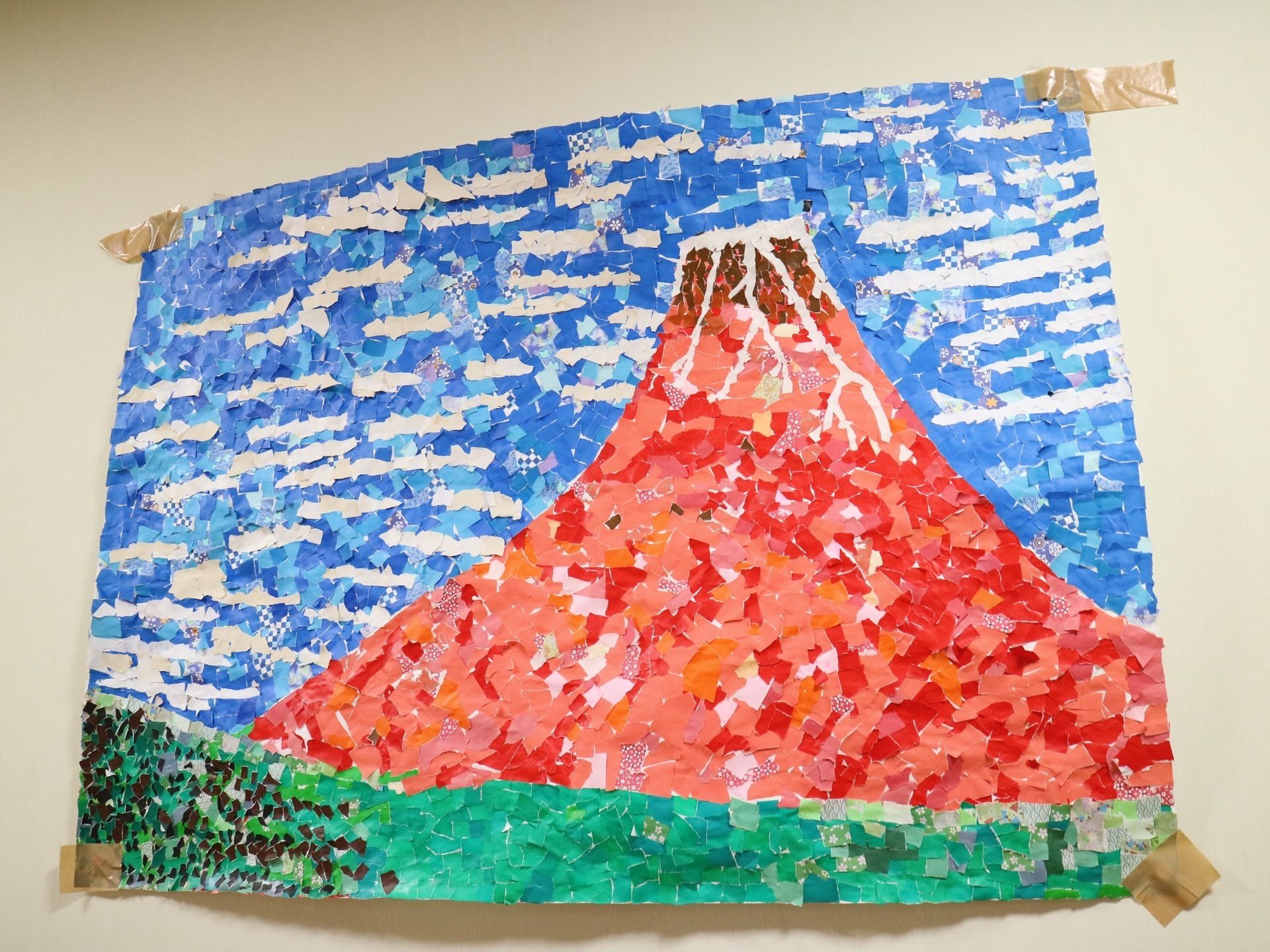 白い壁に貼られた大きな作品の画像である。赤や緑、青い紙をちぎって描かれた赤富士のイラストで、ガムテープで貼られている。