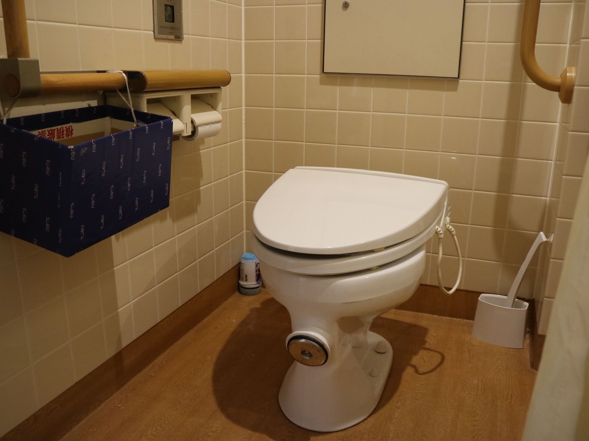ベージュのタイルの壁のトイレで、床はフローリングになっている。中央に洋式便座があり、温水洗浄便座が取り付けられている。