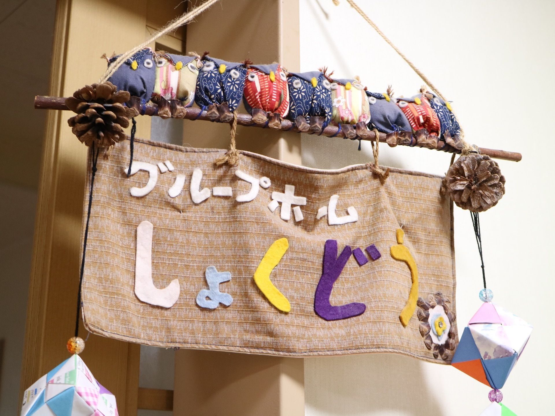 館内に飾られた作品の図である。白い壁の横に引き戸の扉がある。枝や木の実などを使用して作られた看板である。