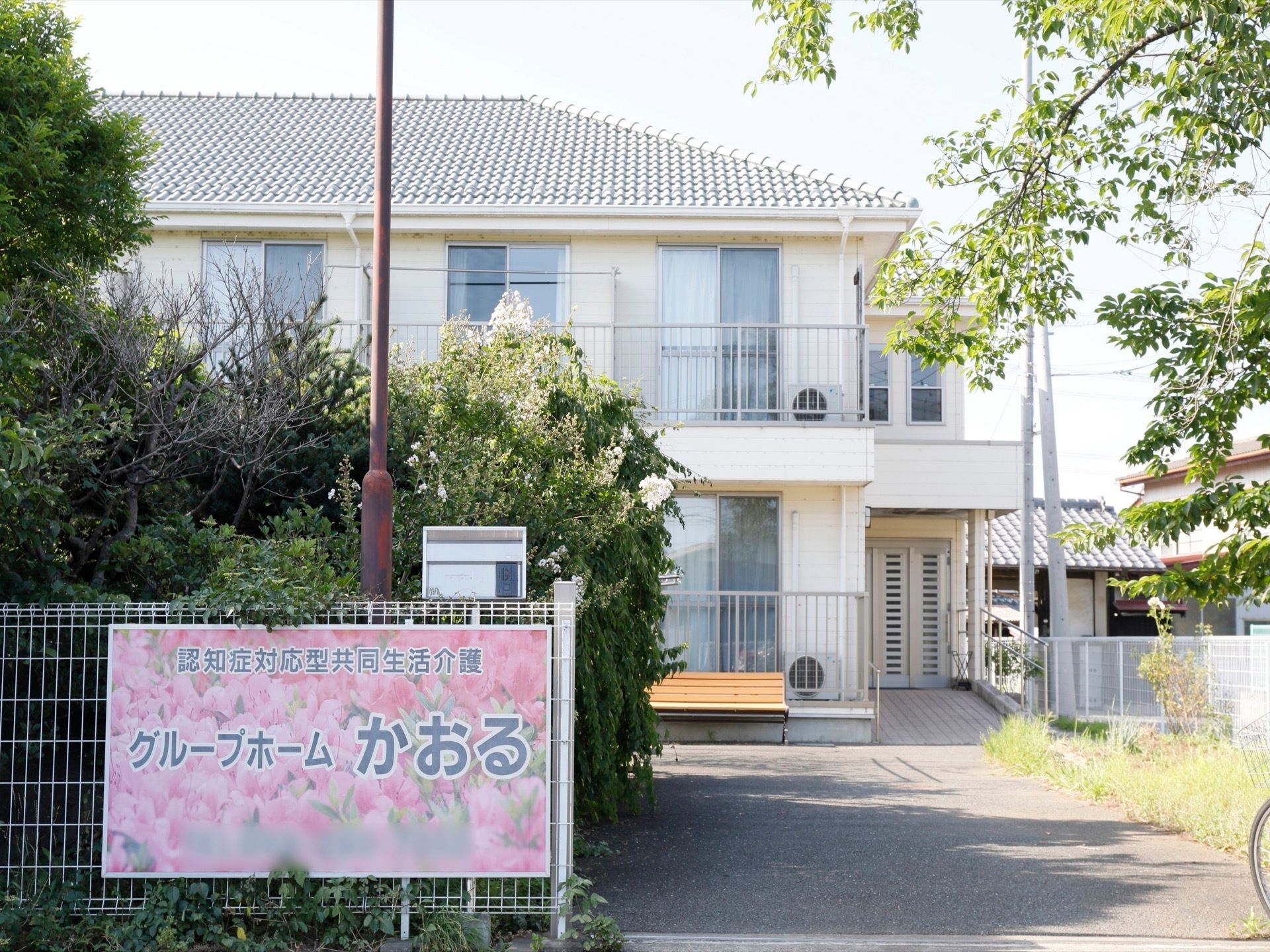 入口の柵に大きな看板が取りつけられており、その上にポストがある。照明が1本立っており、大きな木々が植えられている。
