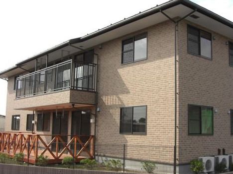 ２階建ての建物。外壁はブラウンのレンガ調。１階にはウッドデッキ、２階にはベランダがある。