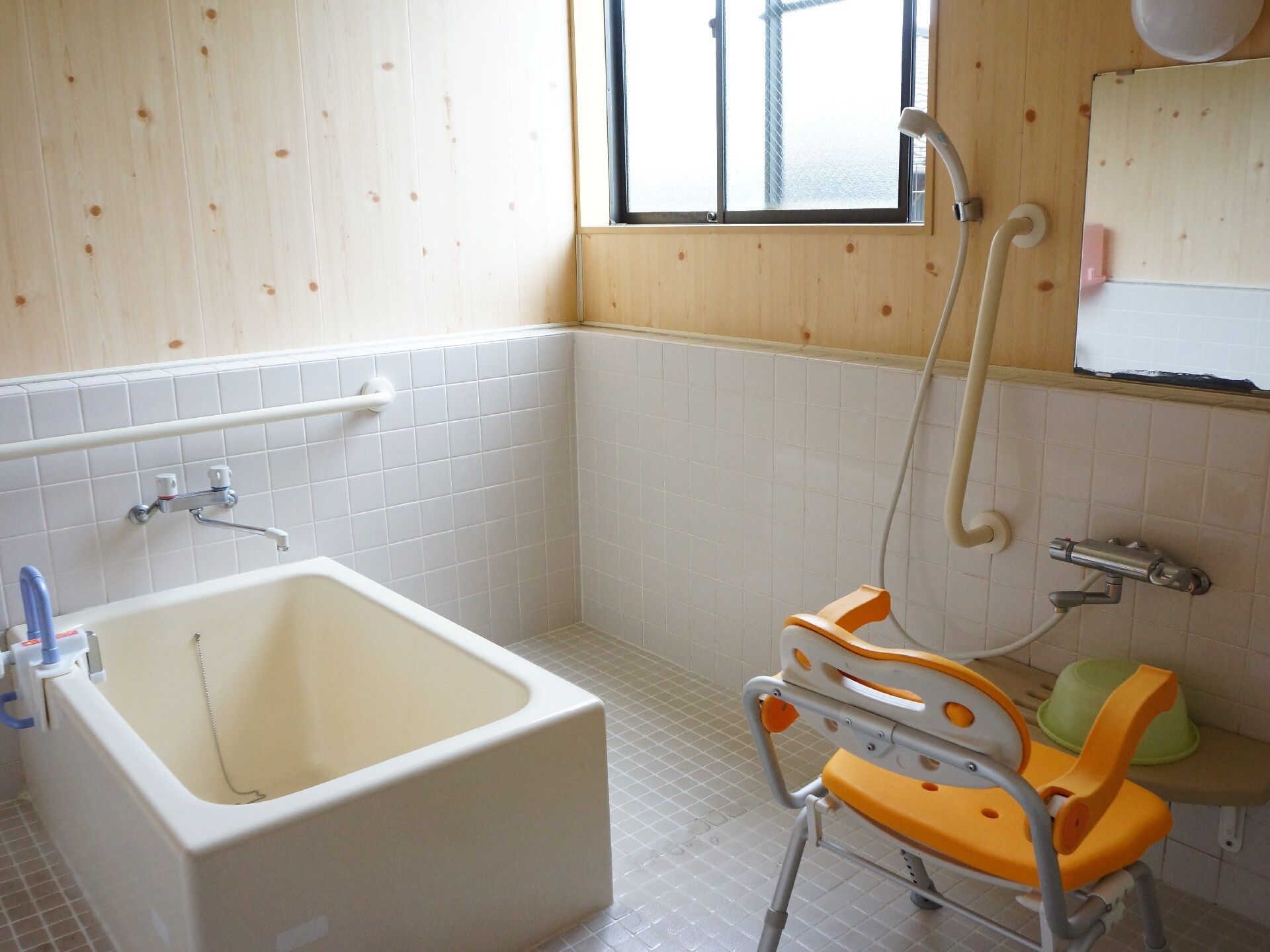 下半分が白いタイル、上半分が木を使用した清潔感あふれる浴室。シャワーチェアや手すりが設置され安全面にも配慮。