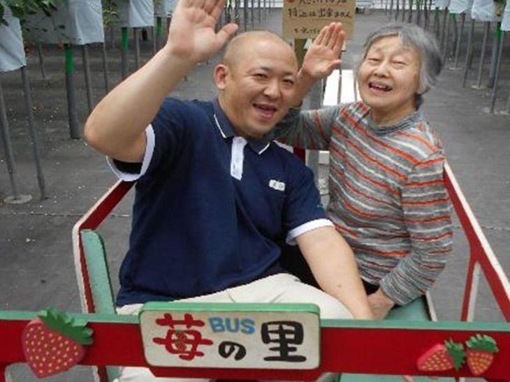 いちご園専用の乗り物に乗り、イチゴ狩りを楽しむ女性入居者と男性スタッフ。二人ともカメラに向かって笑顔で片手を上げている。