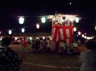 町の納涼祭で行う盆踊り。紅白の幕がかかったやぐらを中心に提灯がつりさげられ、多くの人が踊っている。