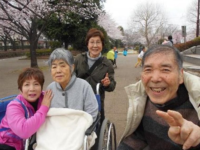 桜並木を笑顔で歩く入居者とスタッフ。男性入居者はピースサイン、車いすに乗った入居者も微笑んでいる。