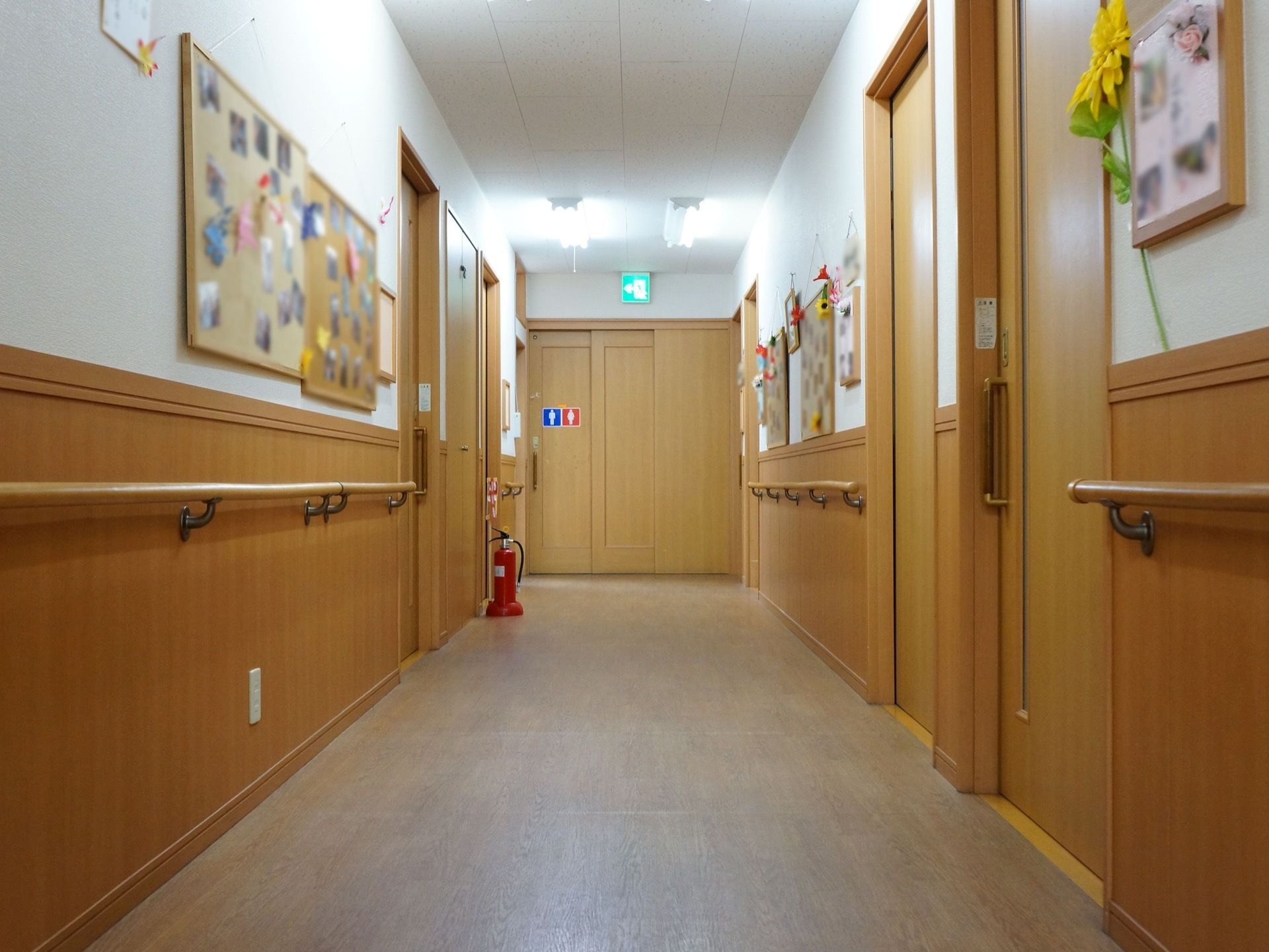 廊下にはコルクボードが多数貼られ、施設でのスナップ写真や折り紙の飾り付けなどが施されている。床に消火器が置かれている。