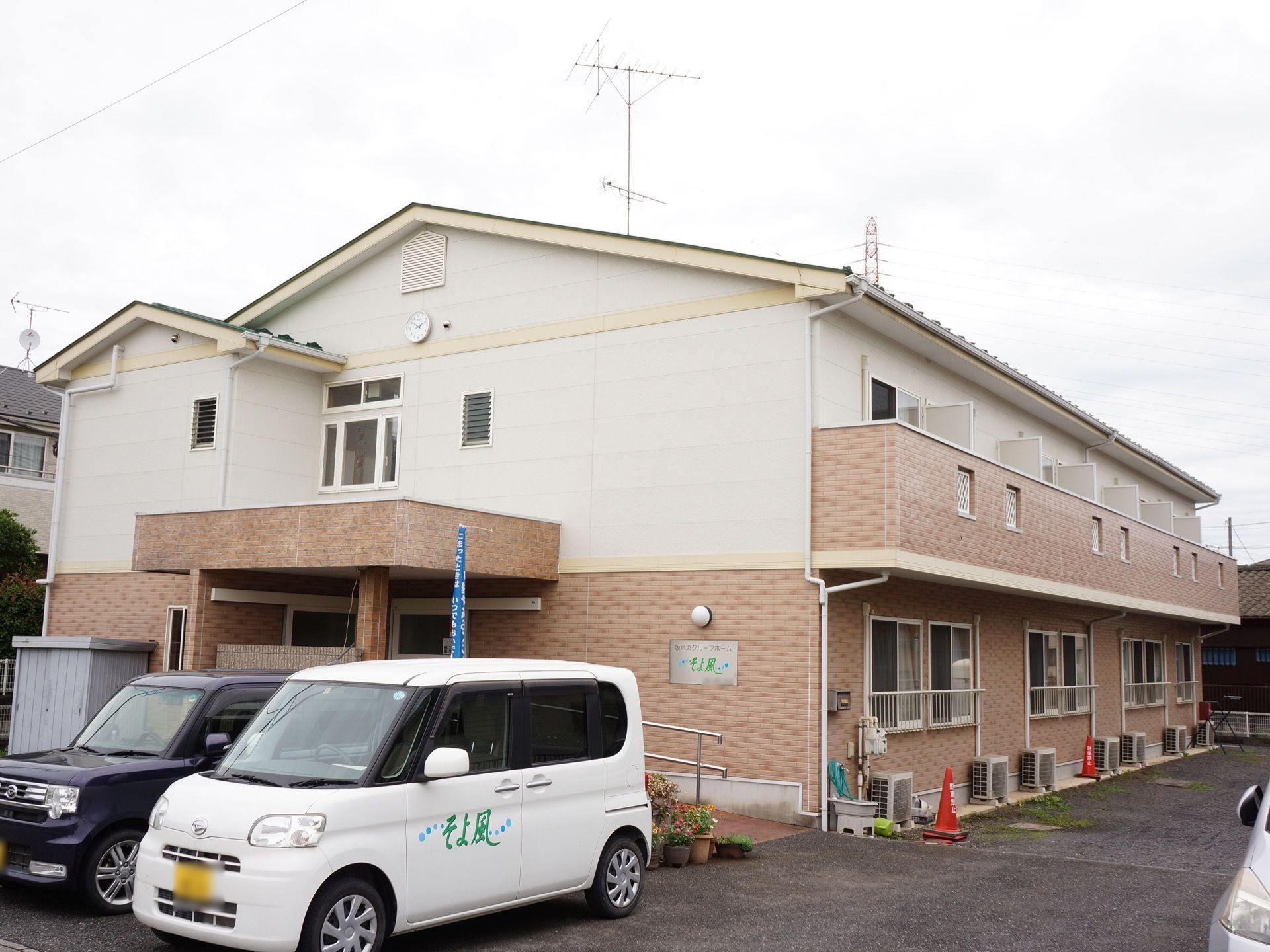白とピンクに塗り分けられた外観になっている。玄関前は駐車スペースになっており、施設の送迎車が停められている。