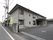 アスファルトで整備された敷地に２階建ての灰色の建物が建っている。道路と敷地の間にはフェンスが設置されている。