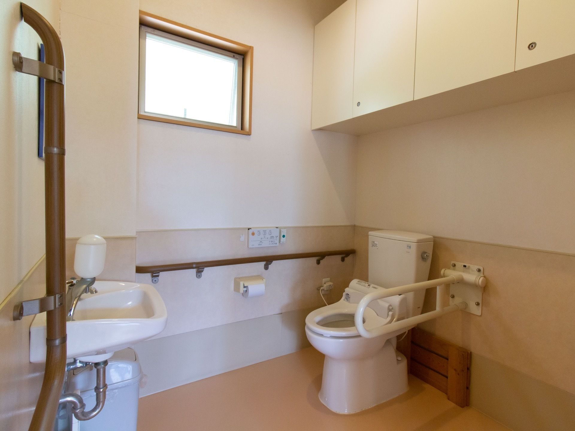 スペースを広く取ったトイレである。手すりや洗面台、呼出ボタンが完備されている。壁には、棚が設置されている。