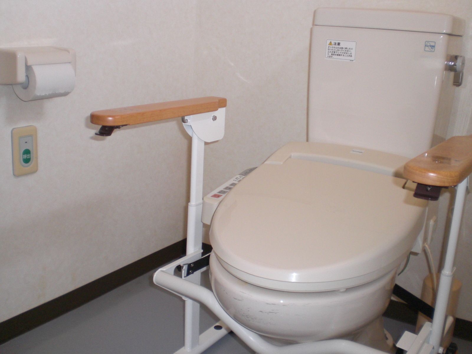 ひじ掛けのついた様式トイレ。壁のトイレットペーパーの下に緊急呼び出し用のブザーがある。