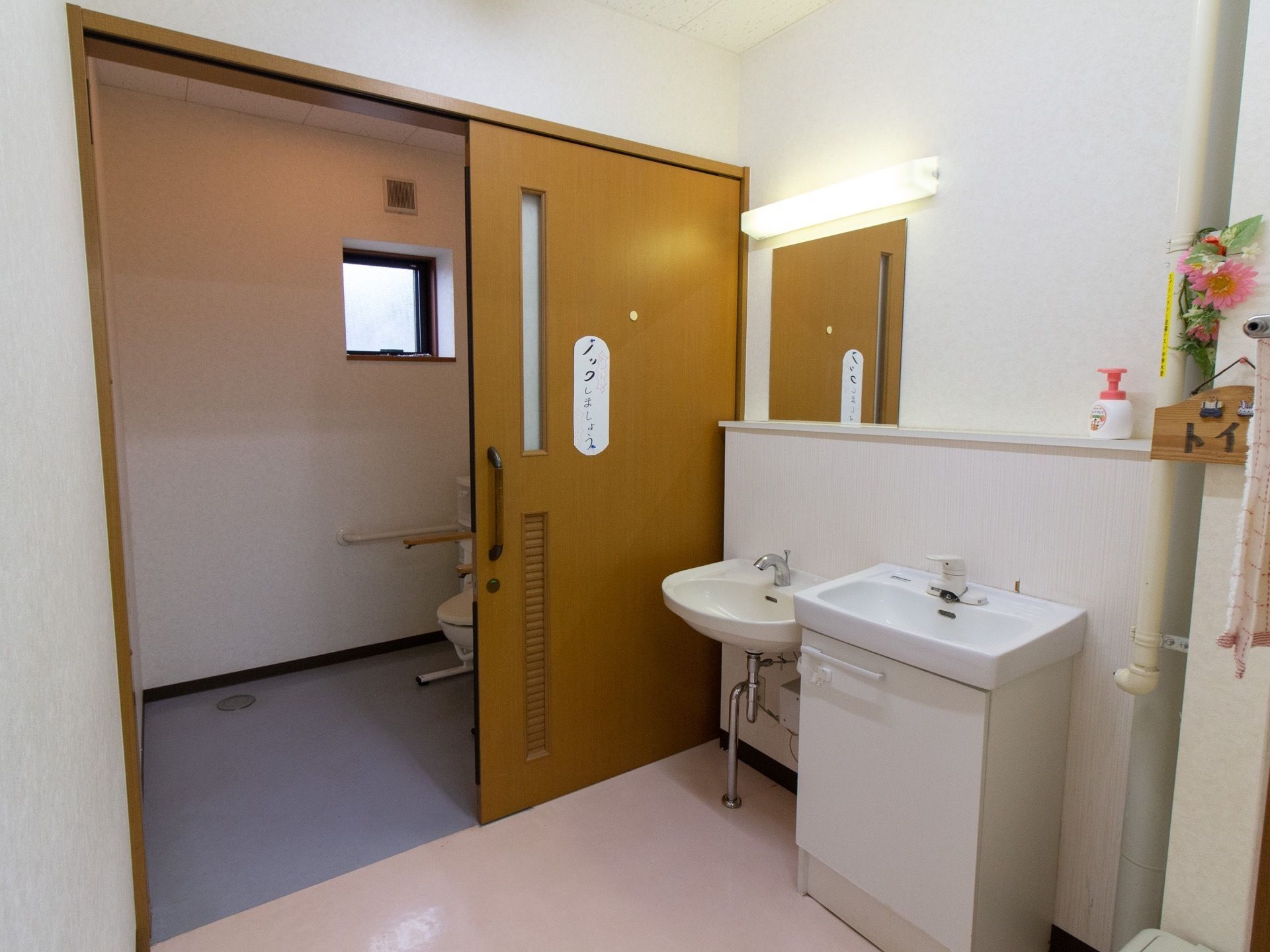 シンクが２つ並んでいる手洗い場の奥に、トイレの個室がある。ドアは引き戸で、便器の両脇に手すりがついているのが見える。