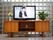 ガラスブロックの前に横長の明るい色の木製のテレビ台に乗った黒い枠のテレビがある。同じ台の上に左に白い花瓶に入った花、右に観葉植物がある。