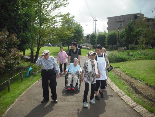 散歩をしているお年寄り、スタッフたち。緑に挟まれた歩道を歩いている。
