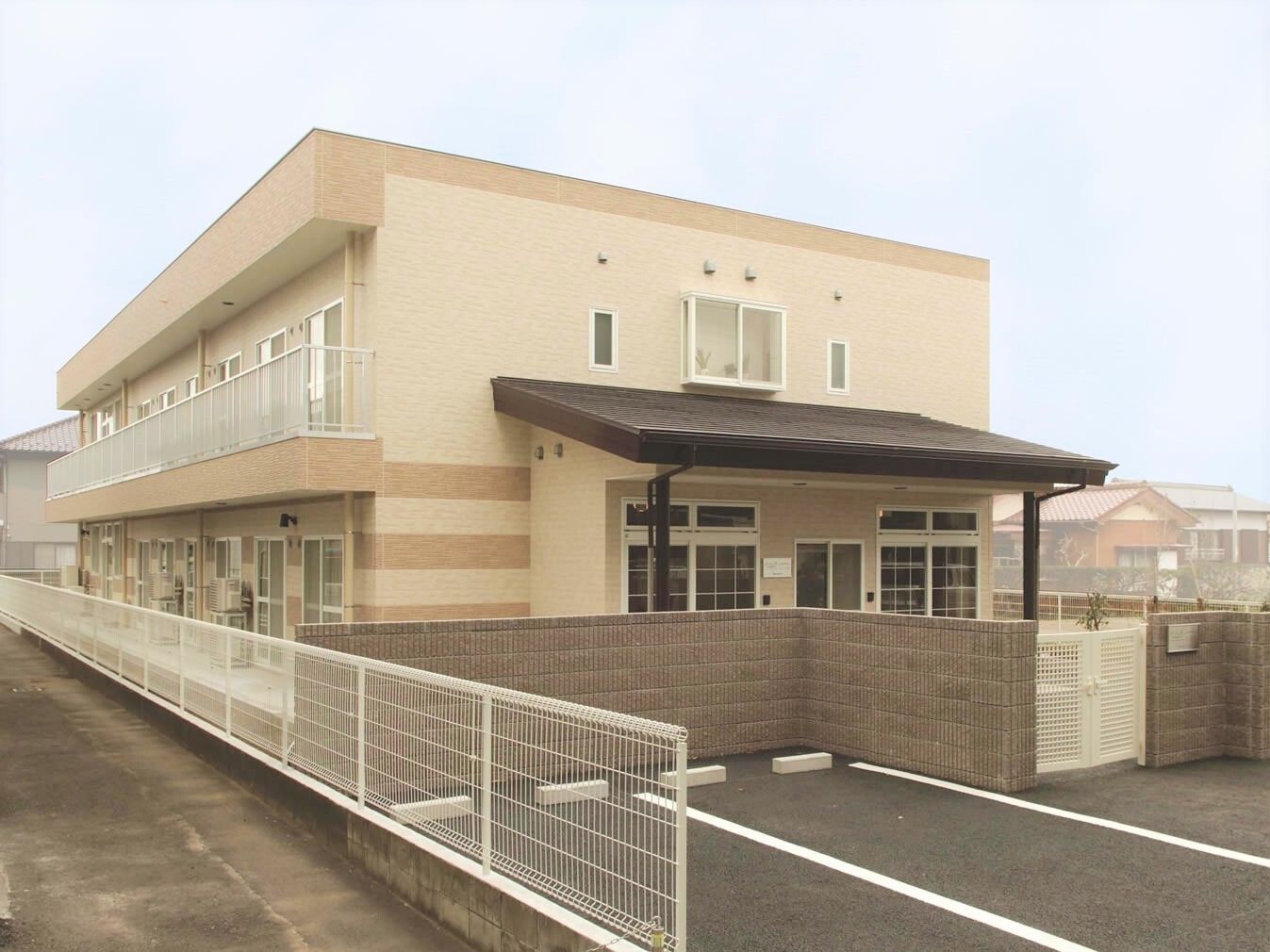 2階建ての建物はベージュ系で温かい雰囲気。建物の玄関には屋根が付き、門の外には駐車スペースも用意されている。周りには住宅も見える。